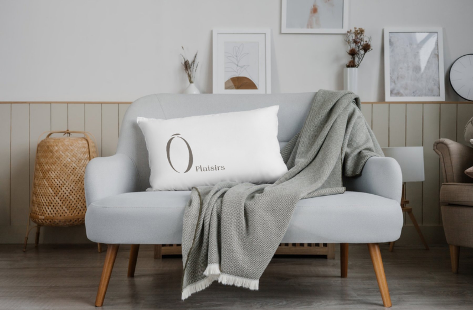Coussin blanc au logo Ô Plaisirs sur un petit divan gris, avec un jeté de laine et des cadres au mur