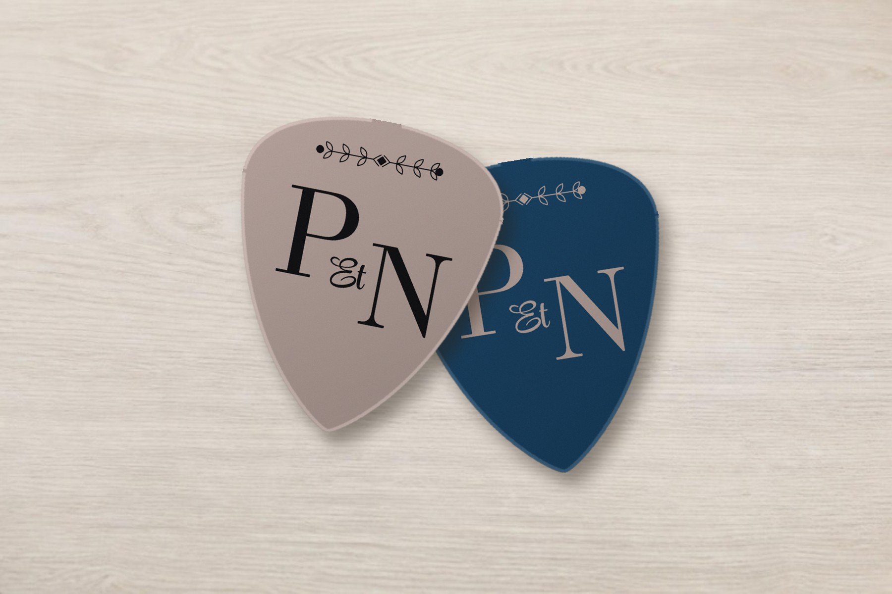 Deux médiators au monogramme P et N posés sur une table en bois beige pâle : l'un taupe avec lettres noires, l'autre bleu marine avec lettres taupe